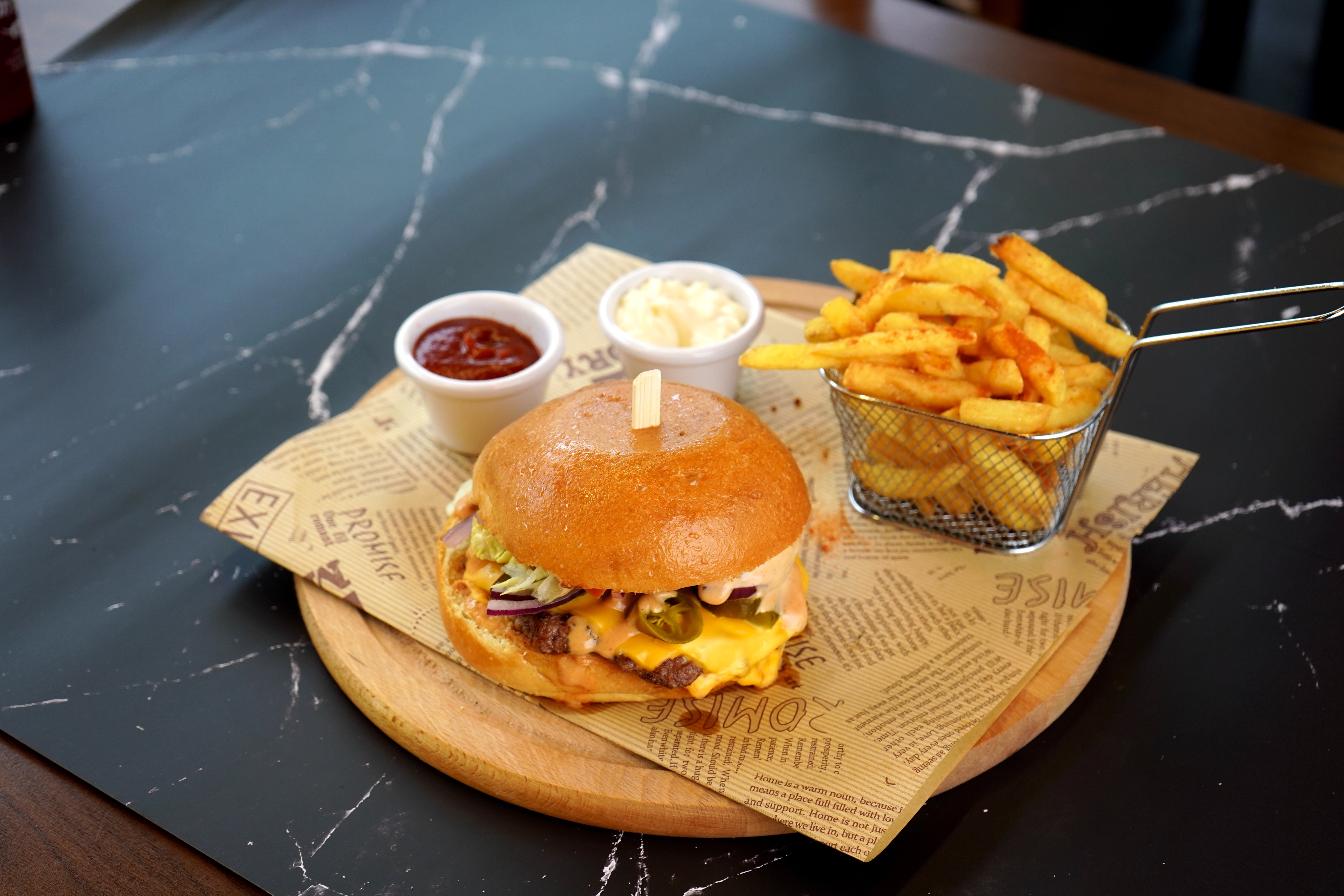 Das Bild zeigt einen frisch zubereiteten Burger mit einer Portion goldbrauner Pommes, serviert mit Ketchup und Mayonnaise im Restaurant The Station Zug. Der Burger besteht aus einem weichen, leicht gerösteten Brötchen, einem saftigen Patty sowie frischen Zutaten wie Salat, Tomaten und einer herzhaften Sauce. Die knusprig frittierten Pommes liegen neben dem Burger und laden zum Genießen ein. Im Hintergrund sind dezente Restaurantdetails von The Station Zug zu erkennen, die eine gemütliche Atmosphäre vermitteln. The Station Zug ist bekannt für seine hochwertigen Zutaten und liebevoll zubereiteten Speisen, was sich in diesem appetitlichen Gericht widerspiegelt.