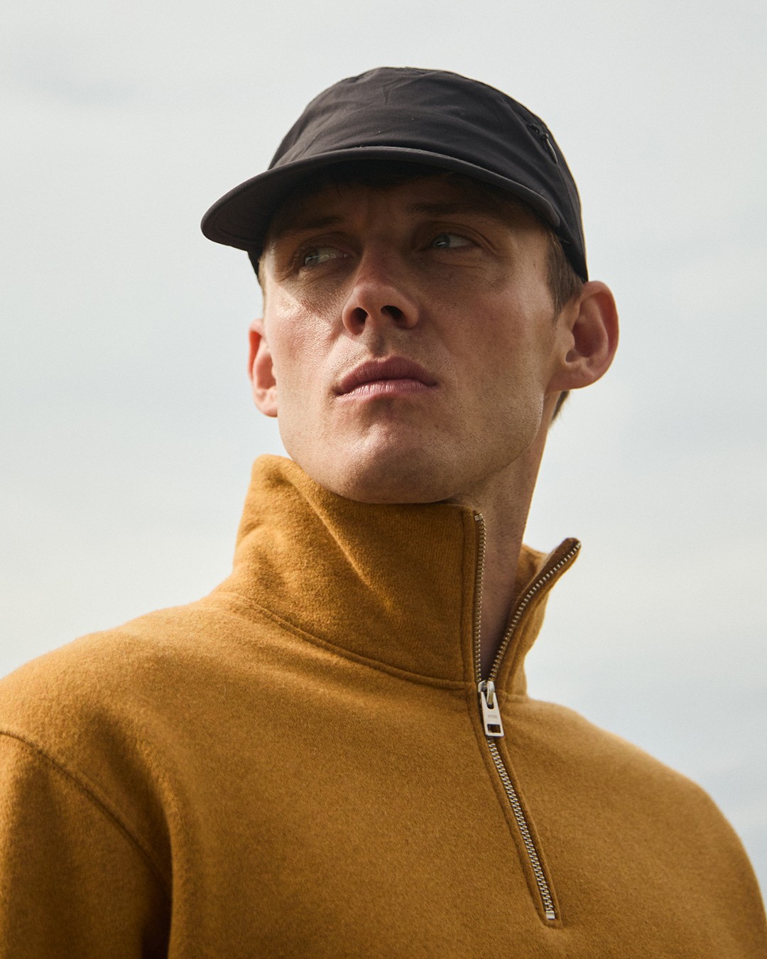 En plan rapproché, un homme regarde au loin portant une casquette noire et un pull col camionneur dans le lookbook de Norse Projects