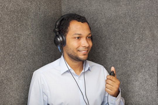 A smiling man wearing headphones holds a microphone, standing against a textured gray background.