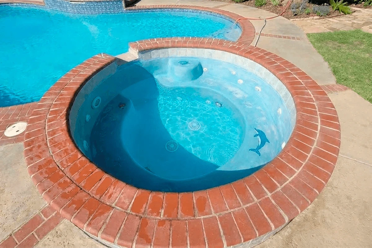 Construcción de piscina residencial moderna con jacuzzi circular integrado, acabados en ladrillo rojo y detalle decorativo de delfín en el fondo.