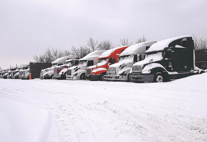 Row of semi-trucks covered in snow parked in a lot during winter.
