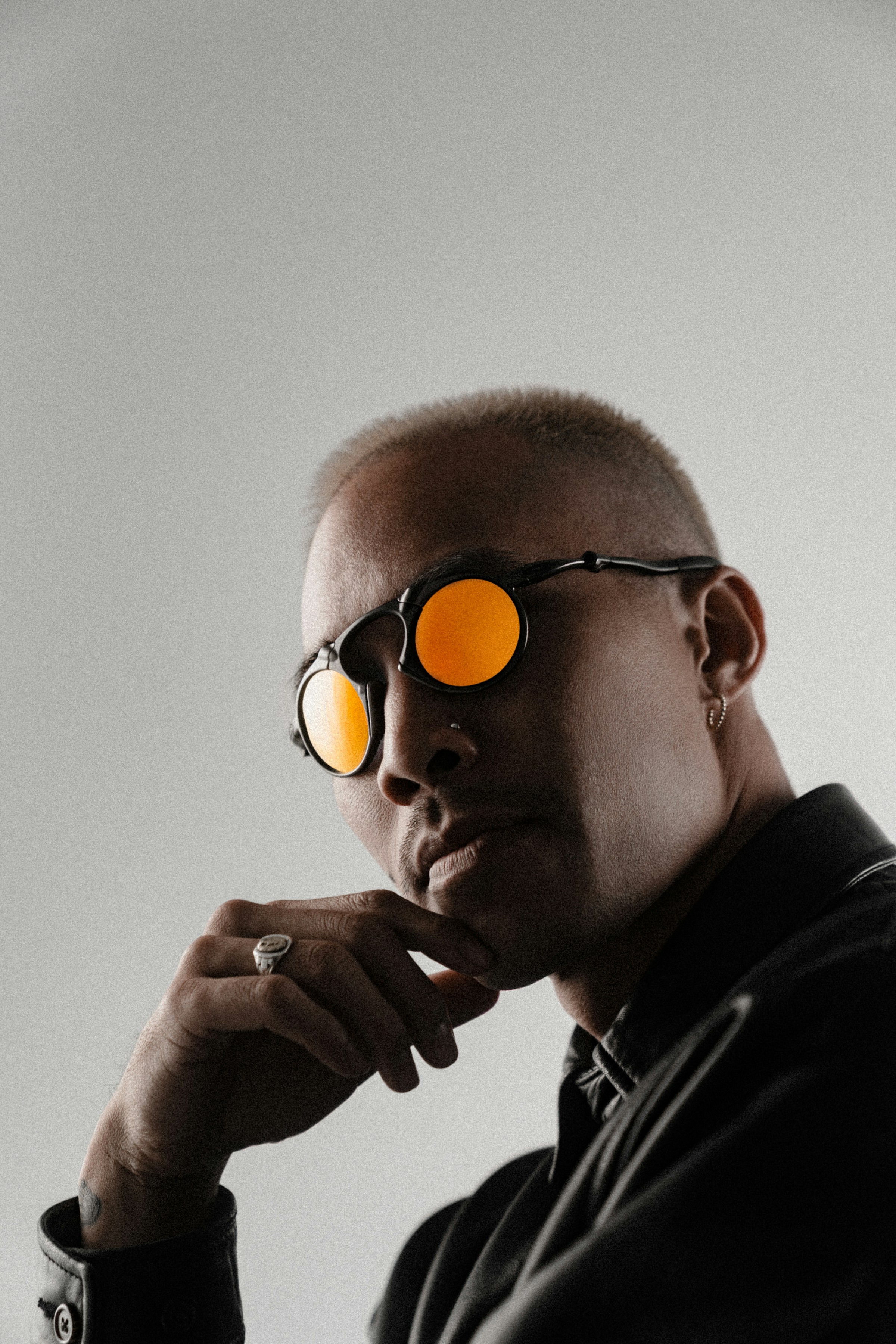 A person wearing stylish, reflective orange sunglasses and a leather jacket poses pensively against a neutral background, with a contemplative expression enhanced by their hand resting on their chin.