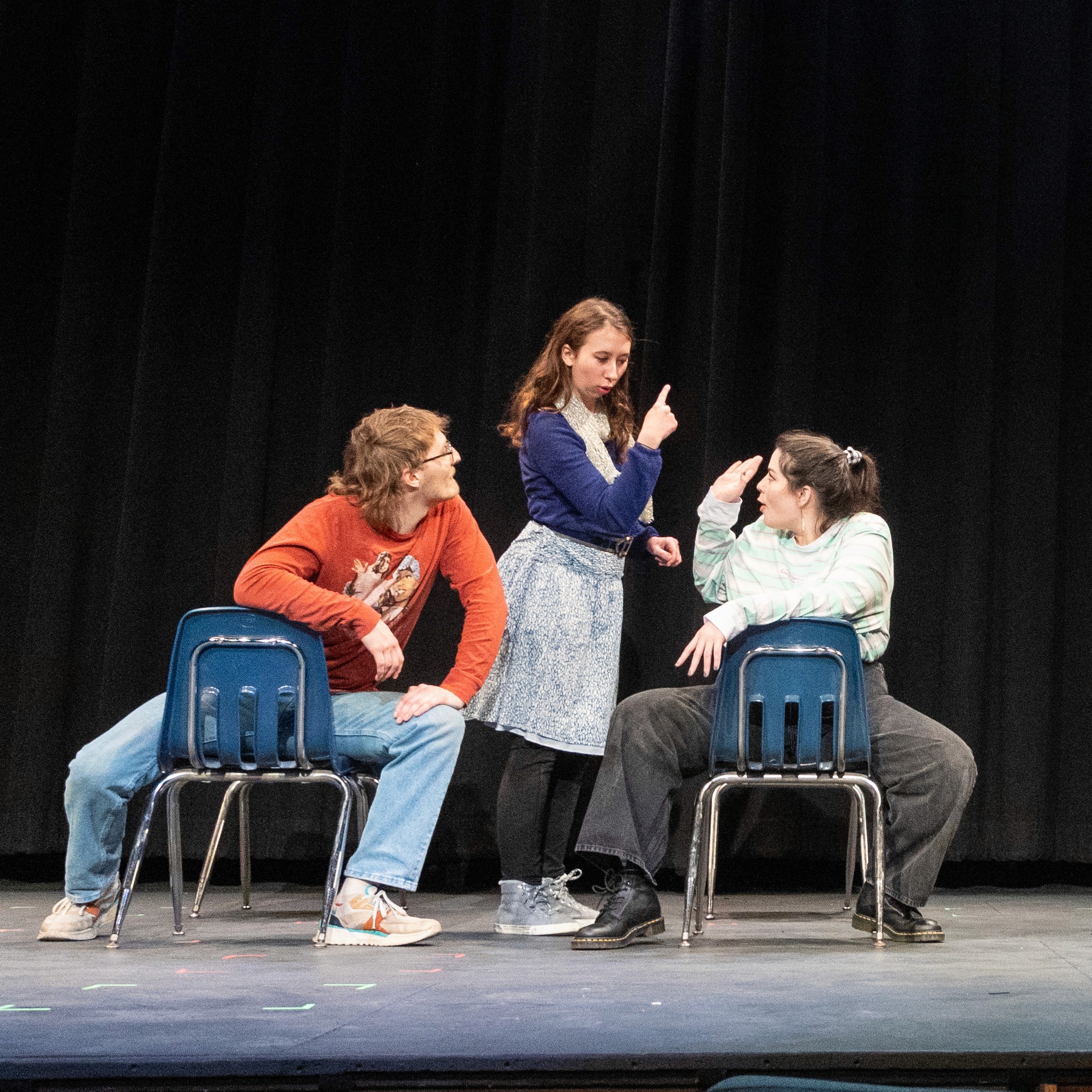 Four theater actors performing a dynamic scene with blue school chairs on a live stage setup.