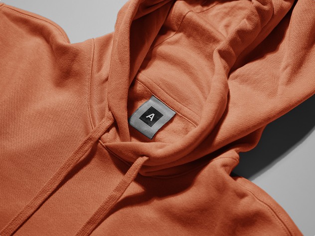 Close-up of an orange hoodie with drawstrings, showing a black inner label with the letter “A.”