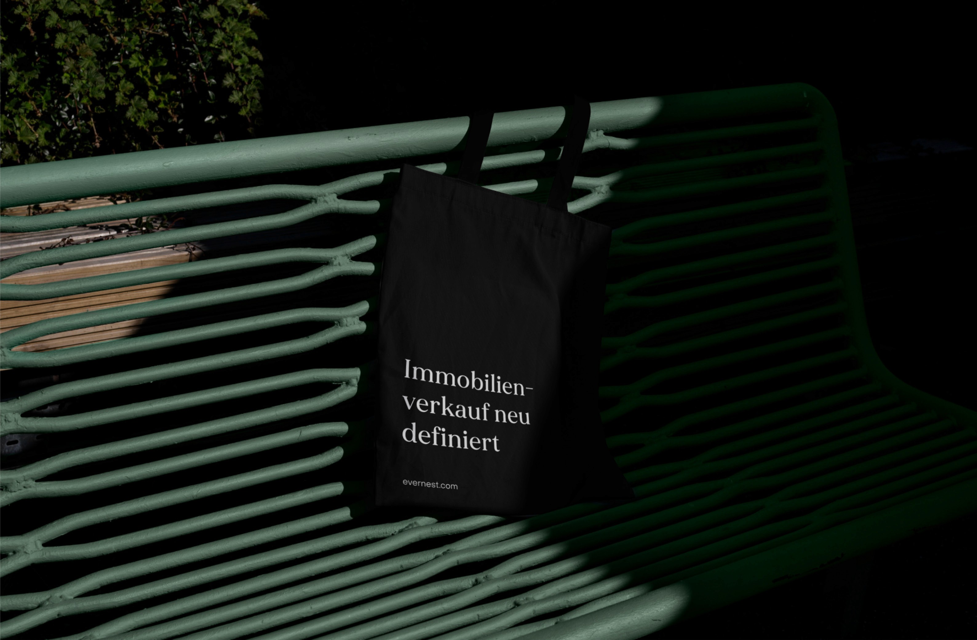 Stylish black tote bag with "Immobilienverkauf neu definiert" text resting on a green bench in a modern urban setting.