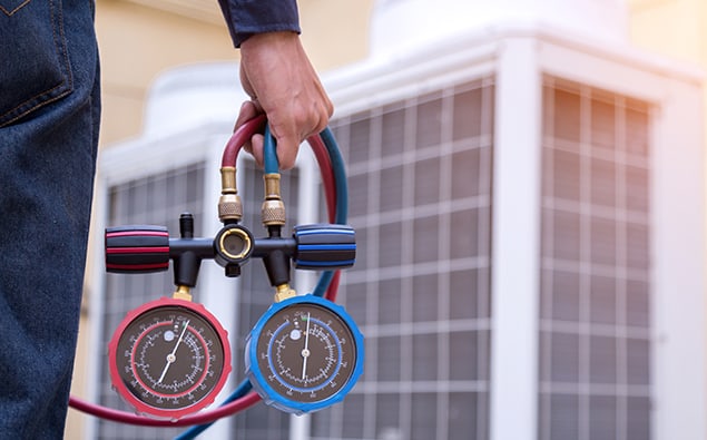 A technician holds a manifold gauge set with red and blue hoses, used for checking and charging HVAC systems. In the background, blurry outdoor air conditioning units can be seen. The scene conveys routine AC repair or maintenance work on air conditioning systems.