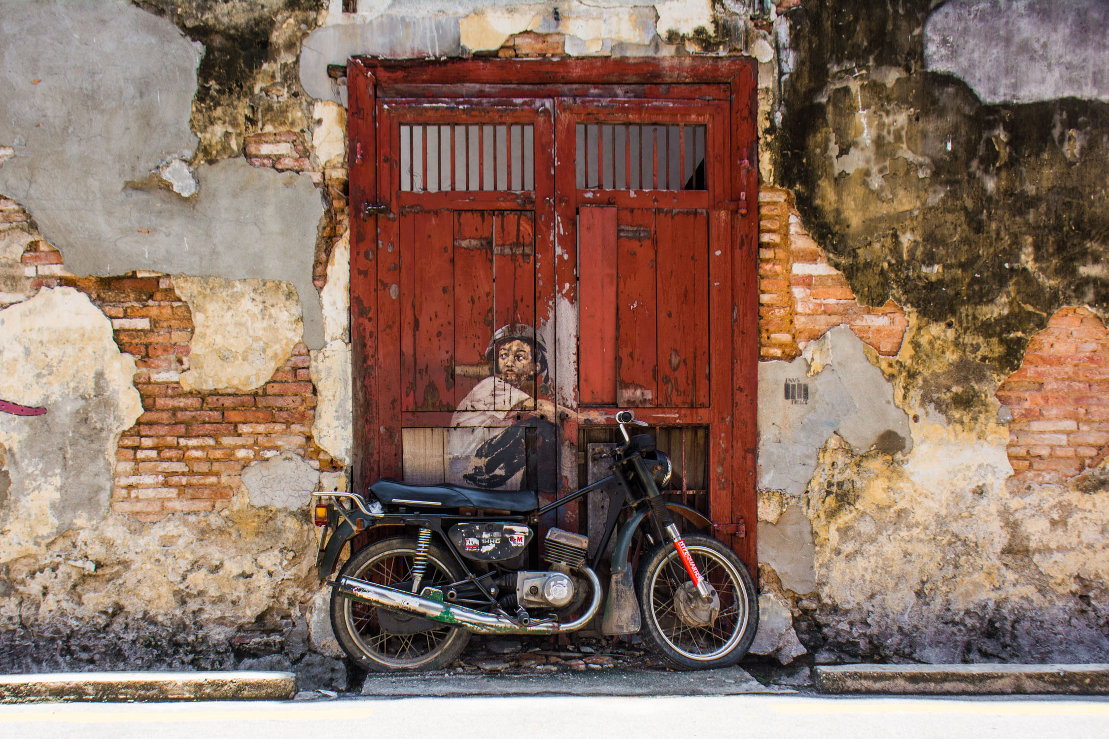 Weathered red wooden doors frame a parked motorcycle against an aged, textured facade bathed in soft daylight.