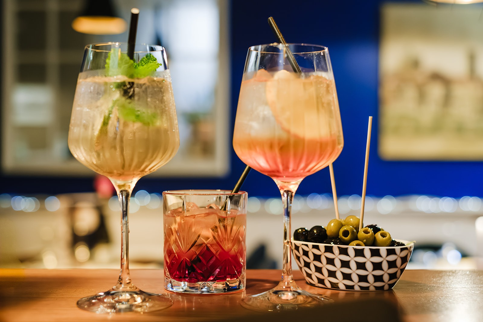 Three drinks on a bar counter: two tall spritz-style cocktails garnished with mint and citrus slices, a shorter glass with an amber-coloured drink, and a small patterned bowl filled with green and black olives on cocktail sticks.
