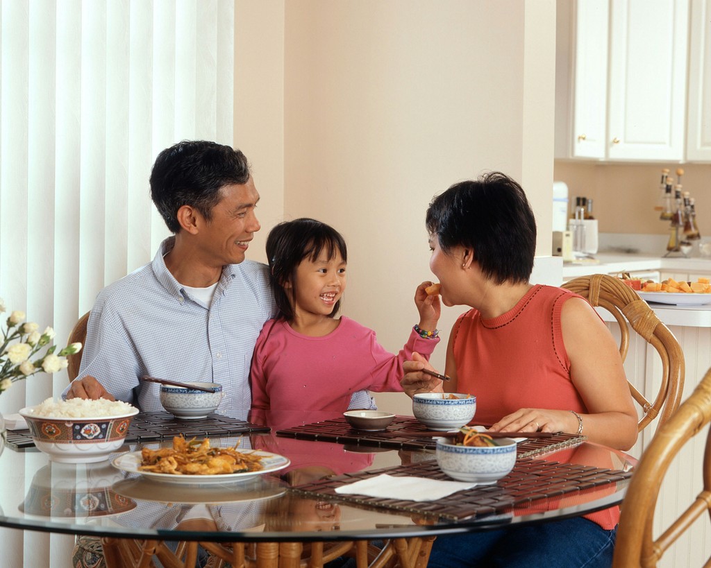 A cheerful Asian family enjoying a meal together at a dining table, with the young daughter playfully feeding her mother, highlighting the warmth and joy of family bonding over food in a cozy home setting.