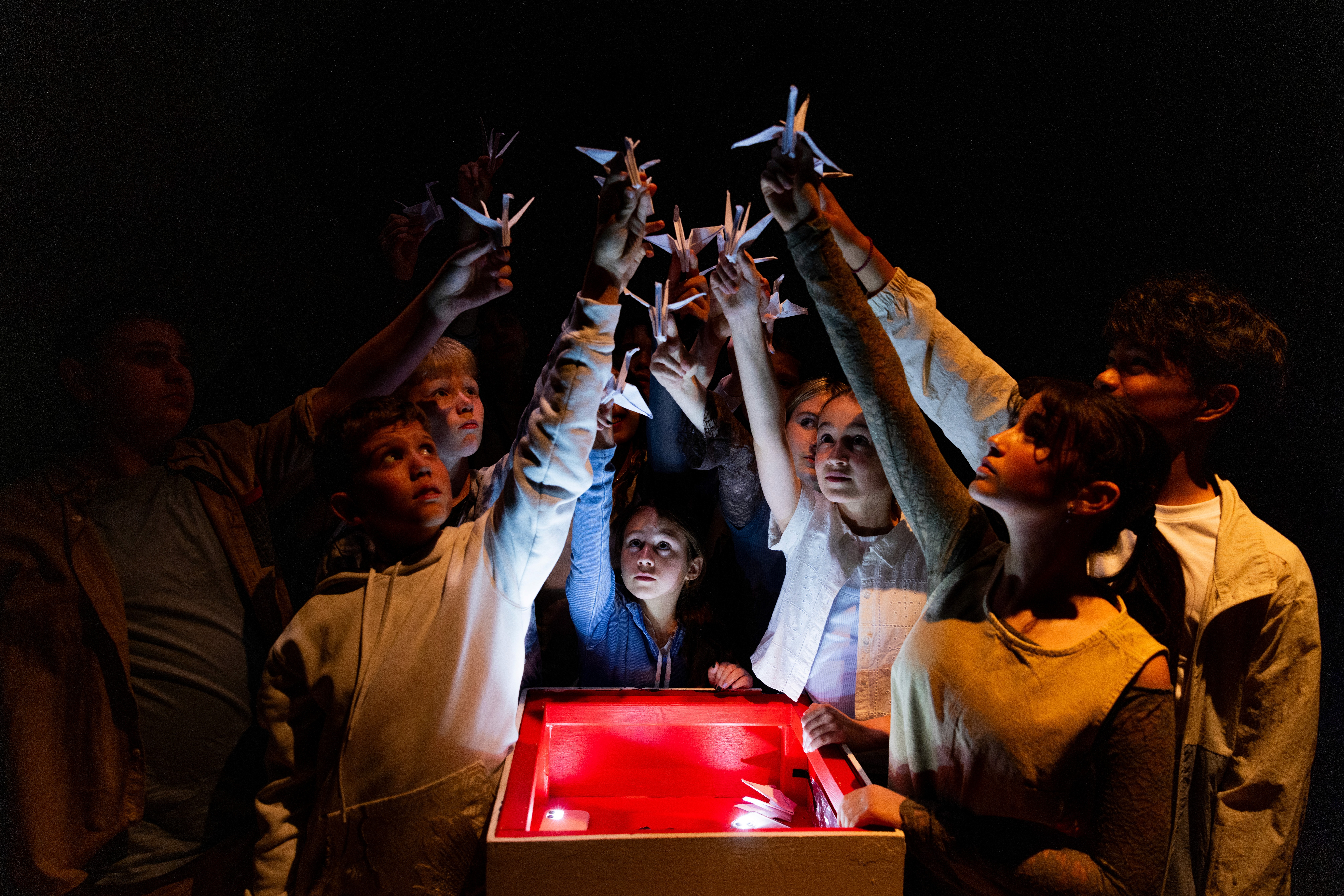 A group of young performers raise paper cranes into the light with a glowing red box beneath them, their faces illuminated with reverence in a darkened theatre.