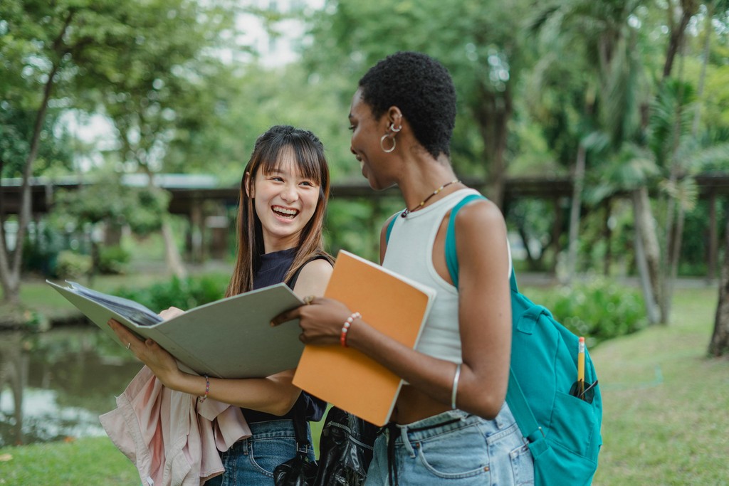 Two international students carrying notebooks and a backpack, smiling and conversing while walking outdoors on a campus.