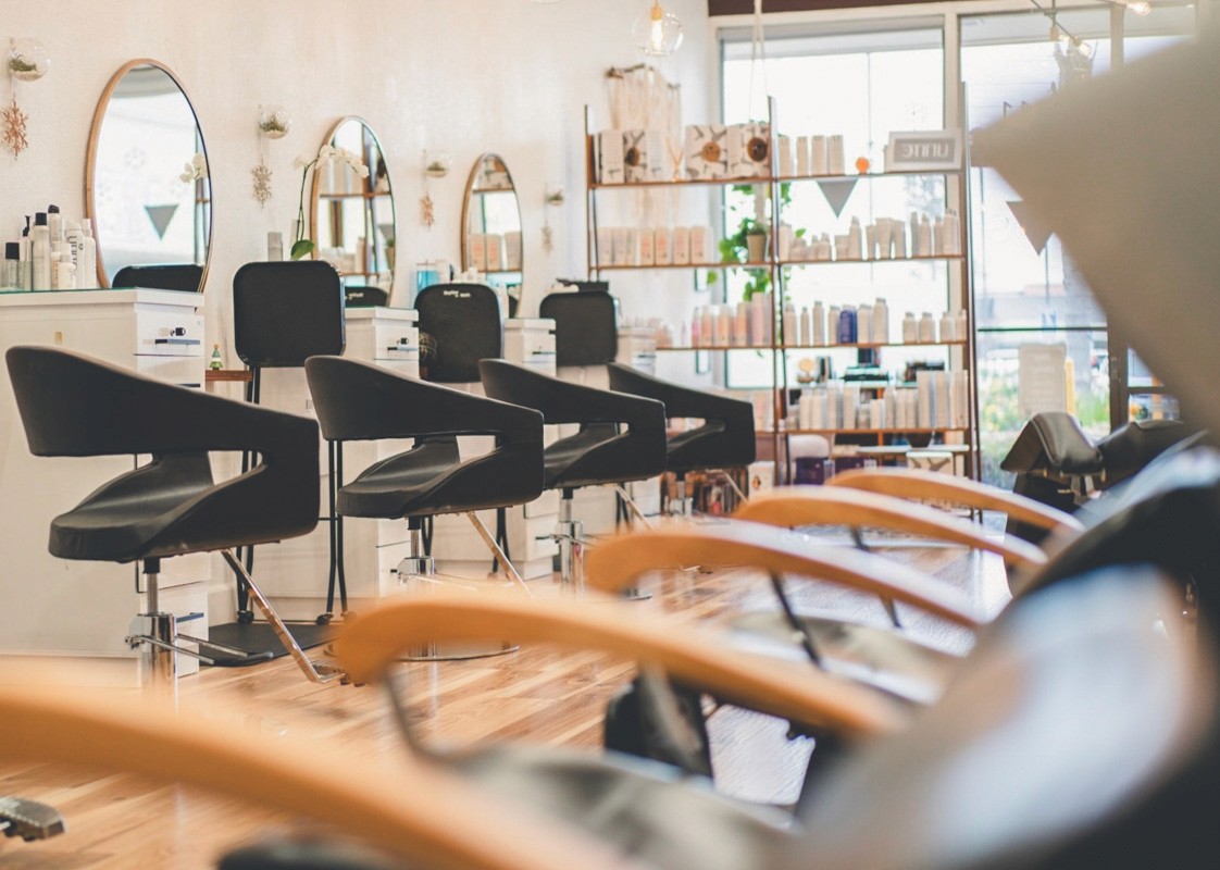 View of Del Sol Hair Studio styling stations from the shampoo bowl area, showcasing black chairs, round mirrors, and natural light filtering through the front windows.