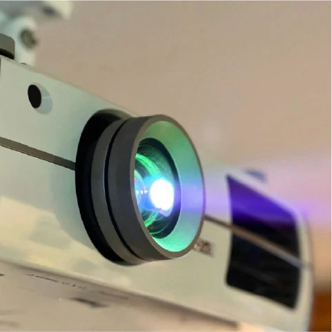 Close-up of a projector lens emitting a bright light, casting a greenish hue. The projector is mounted on a ceiling, ready for a presentation.