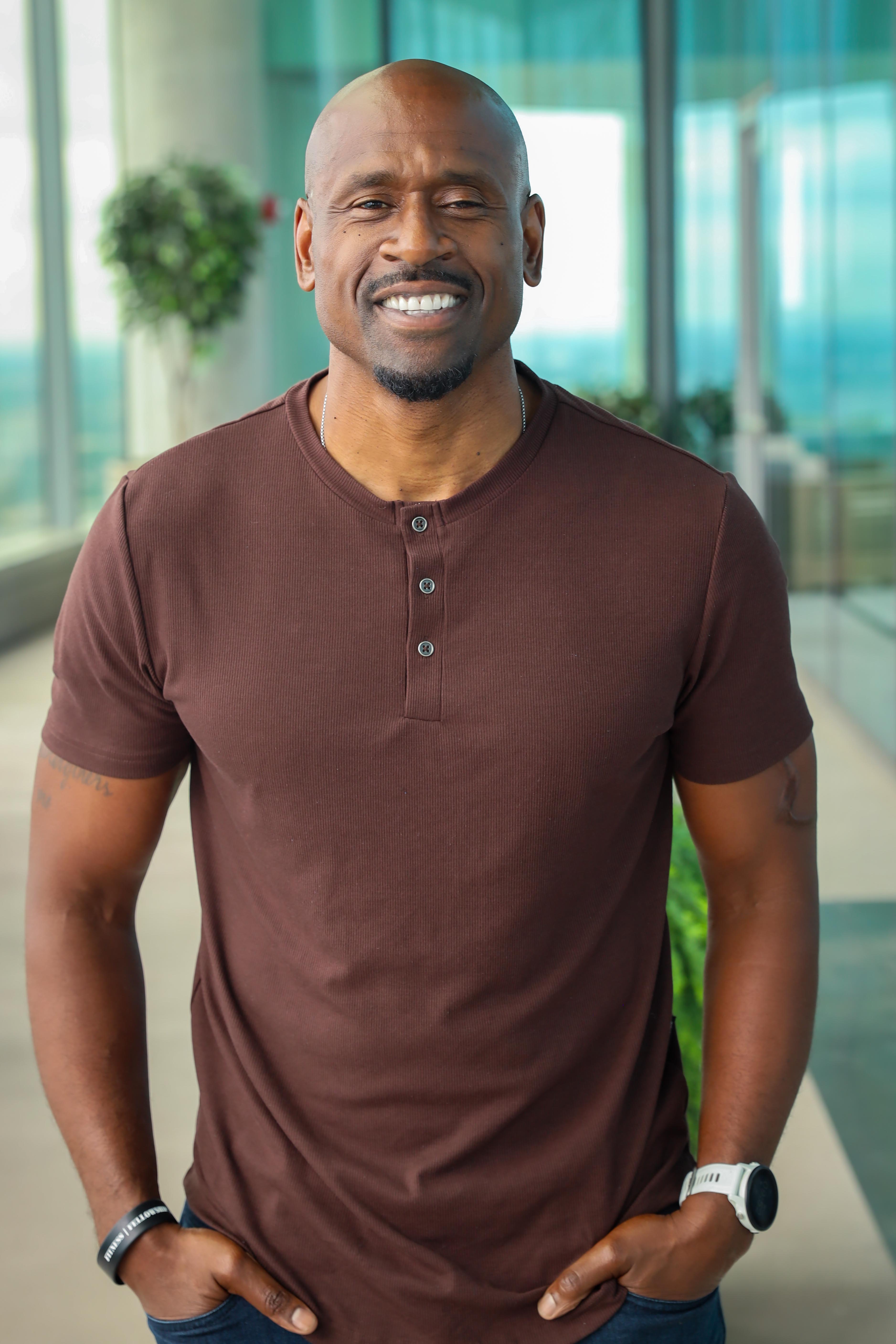 A smiling person with wavy blonde hair, wearing a black top, posed against a warm brown background.