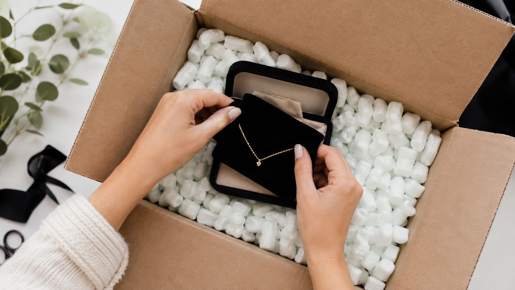 A person carefully packages a delicate gold necklace in a velvet jewelry box within a shipping container.