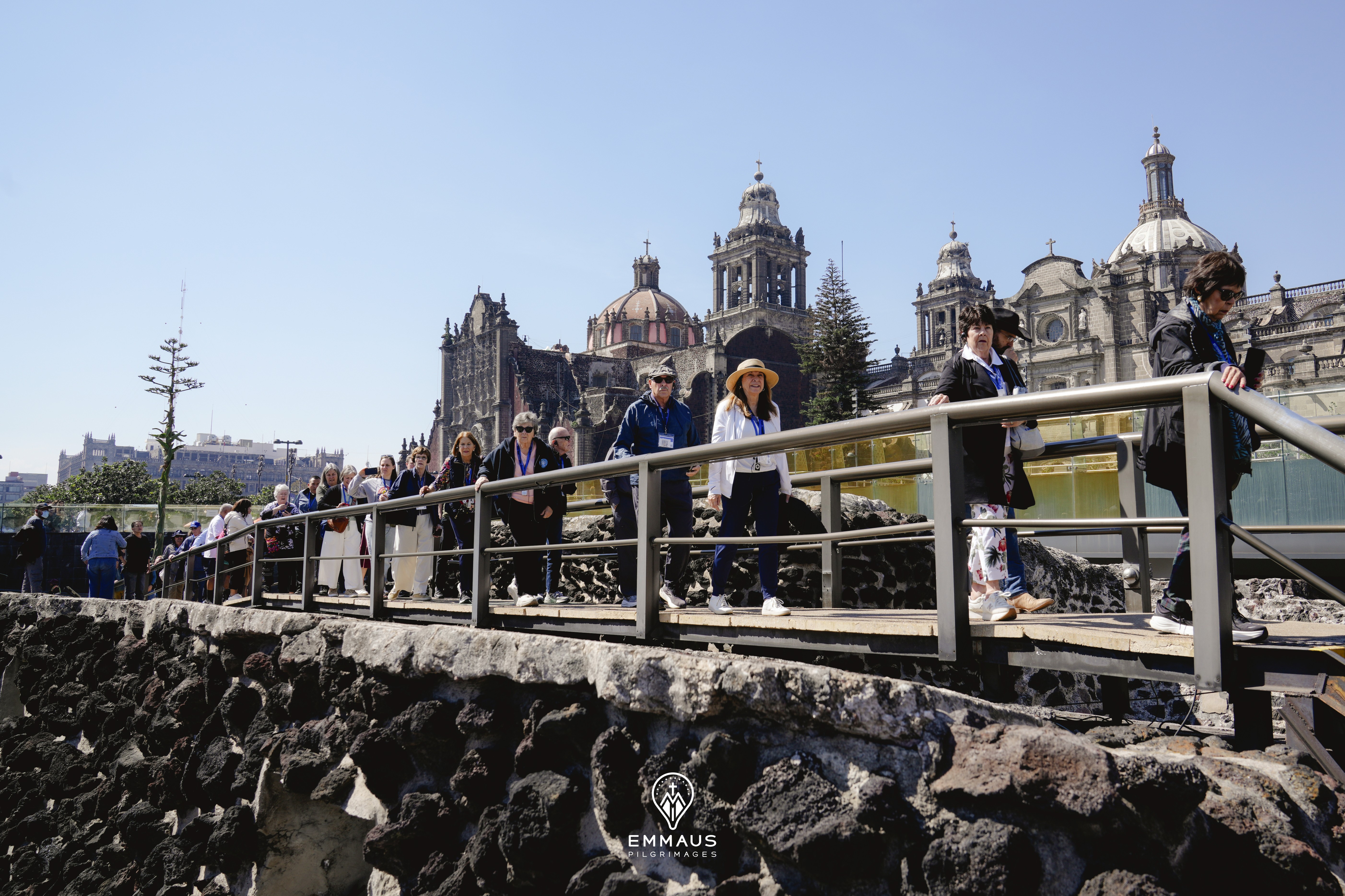Tourists walking across a stone bridge towards the Metropolitan Cathedral in Mexico City on a sunny day.