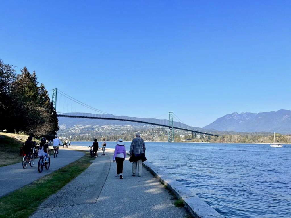 Sunny day with blue skies and the Lion's Gate bridge connecting Stanley Park to North Vancouver. A few kids are cycling and an elderly couple is walking alonside.
