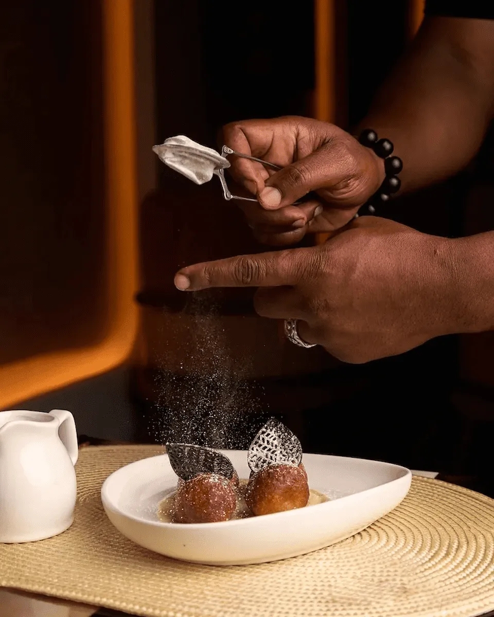 A chef dusts powdered sugar over a gourmet bofrot.