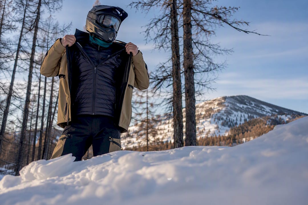 Snowmobile rider adjusting an outer layer in a snowy mountain landscape