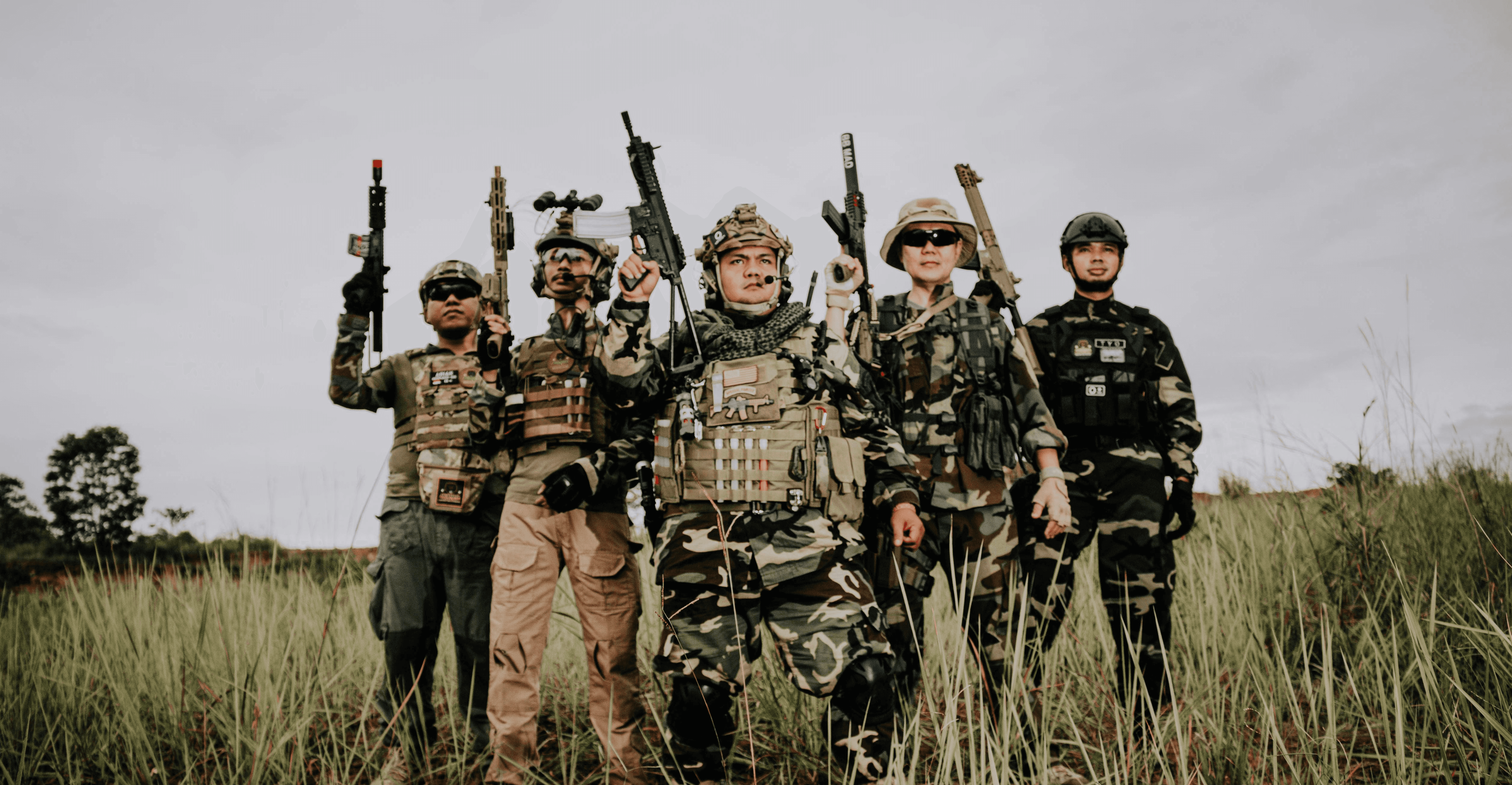 Five soldiers in the foreground holding weapons in their hands, with a battle-damaged field in the background.