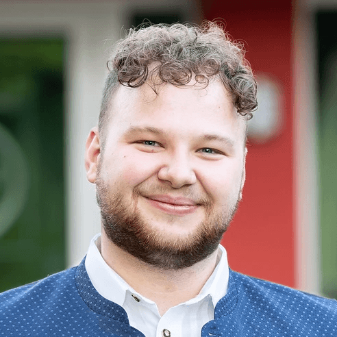 Tobias Huber ein junger Mann mit Bart sthet vor einer Wand und lächelt leicht