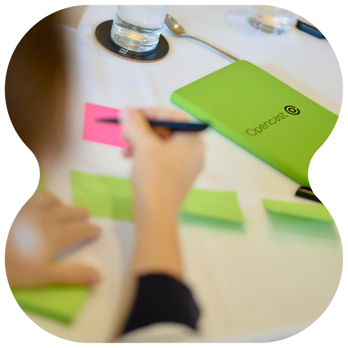 Hand writing on colorful sticky notes at a table with a green Opencast notebook, glass of water, and spoon nearby.