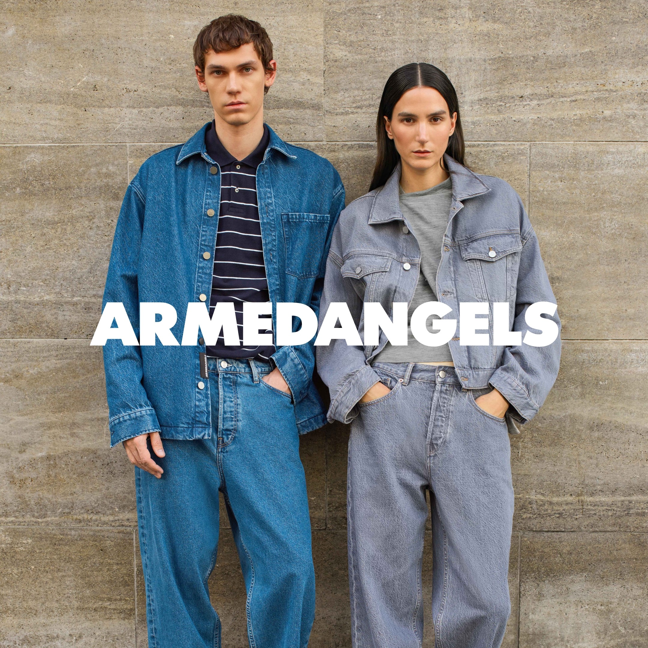Armedangels denim - a man and a woman lean against a grey wall wearing denim outfits