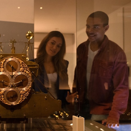 يبتسم شخصان أثناء النظر إلى ساعة أثرية قديمة متقنة الصنع داخل صندوق عرض في متحف.