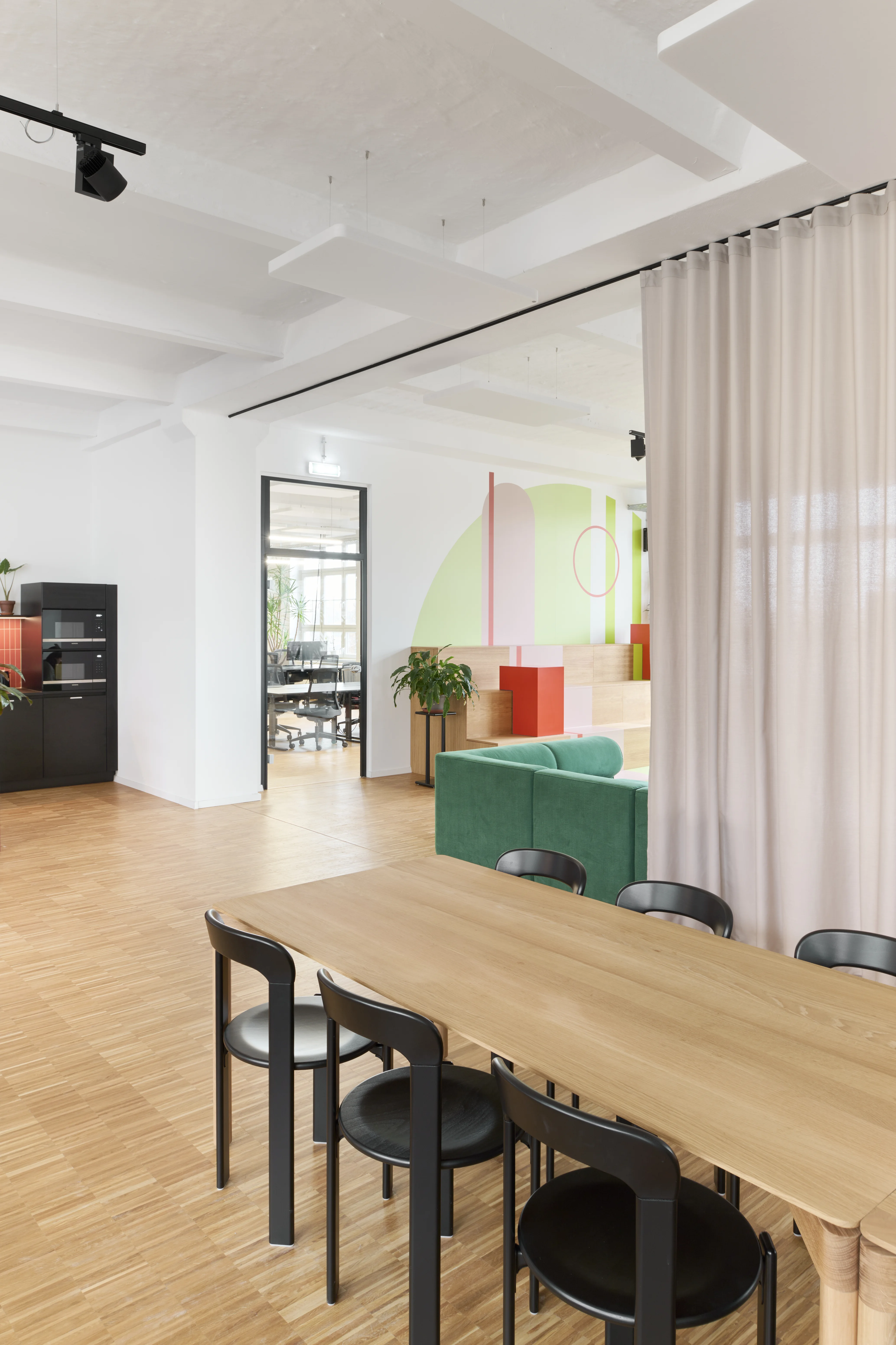 Dining area at PLAN A with an oak table and black chairs, a beige room-height curtain partition, a green sofa and a playful seating landscape with pastel wall graphics in the background.