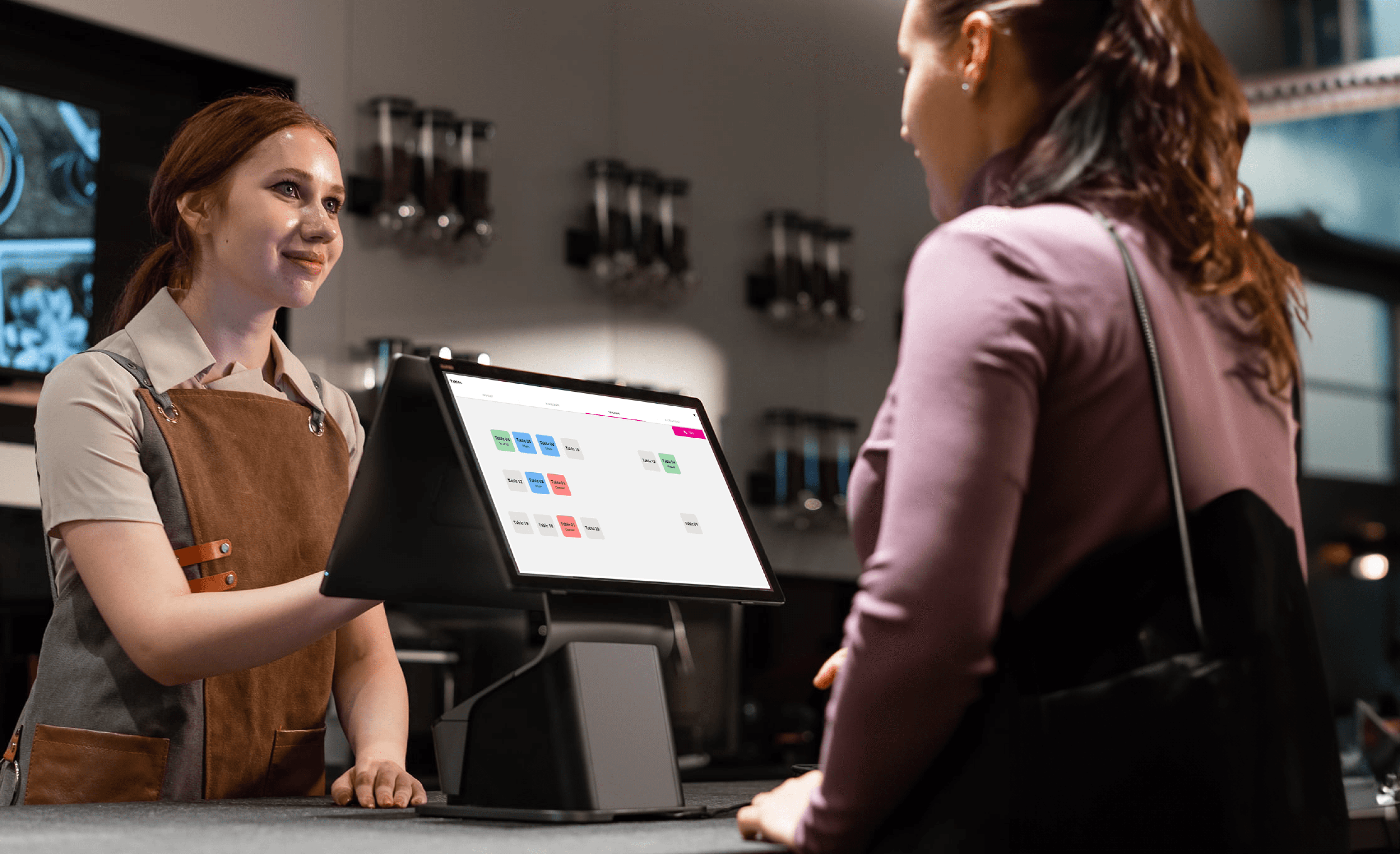 A young person with curly hair smiles at the camera while sitting in front of a laptop against a dark background.