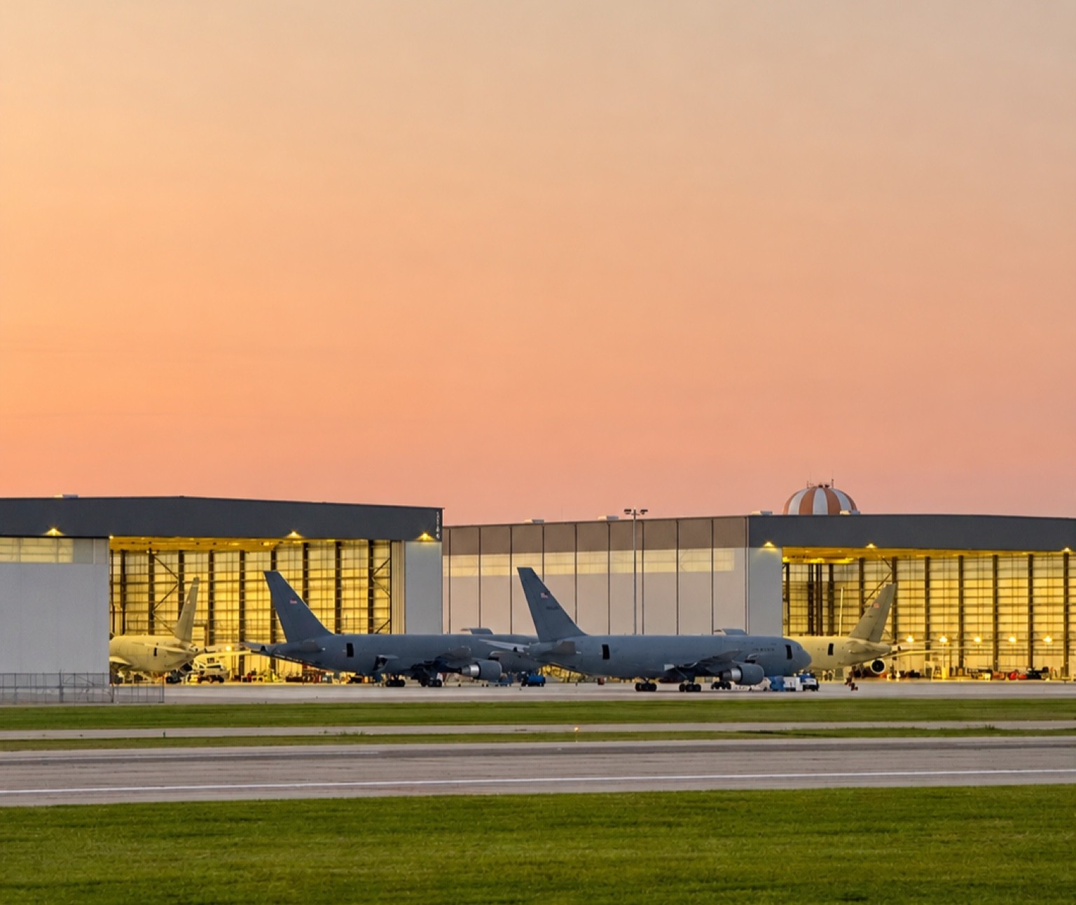 Two KC-46A Pegasus tankers parked outside brightly lit hangars at dusk