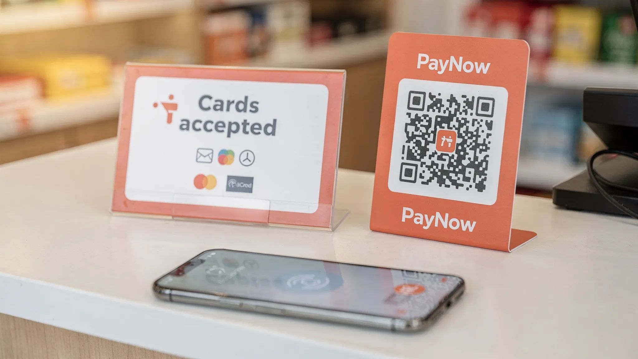 A small Singapore retail checkout counter with a clean card payment sign, PayNow QR display, and a smartphone ready for Tap-to-Phone payment. The sign clearly says cards accepted and shows simple payment icons.