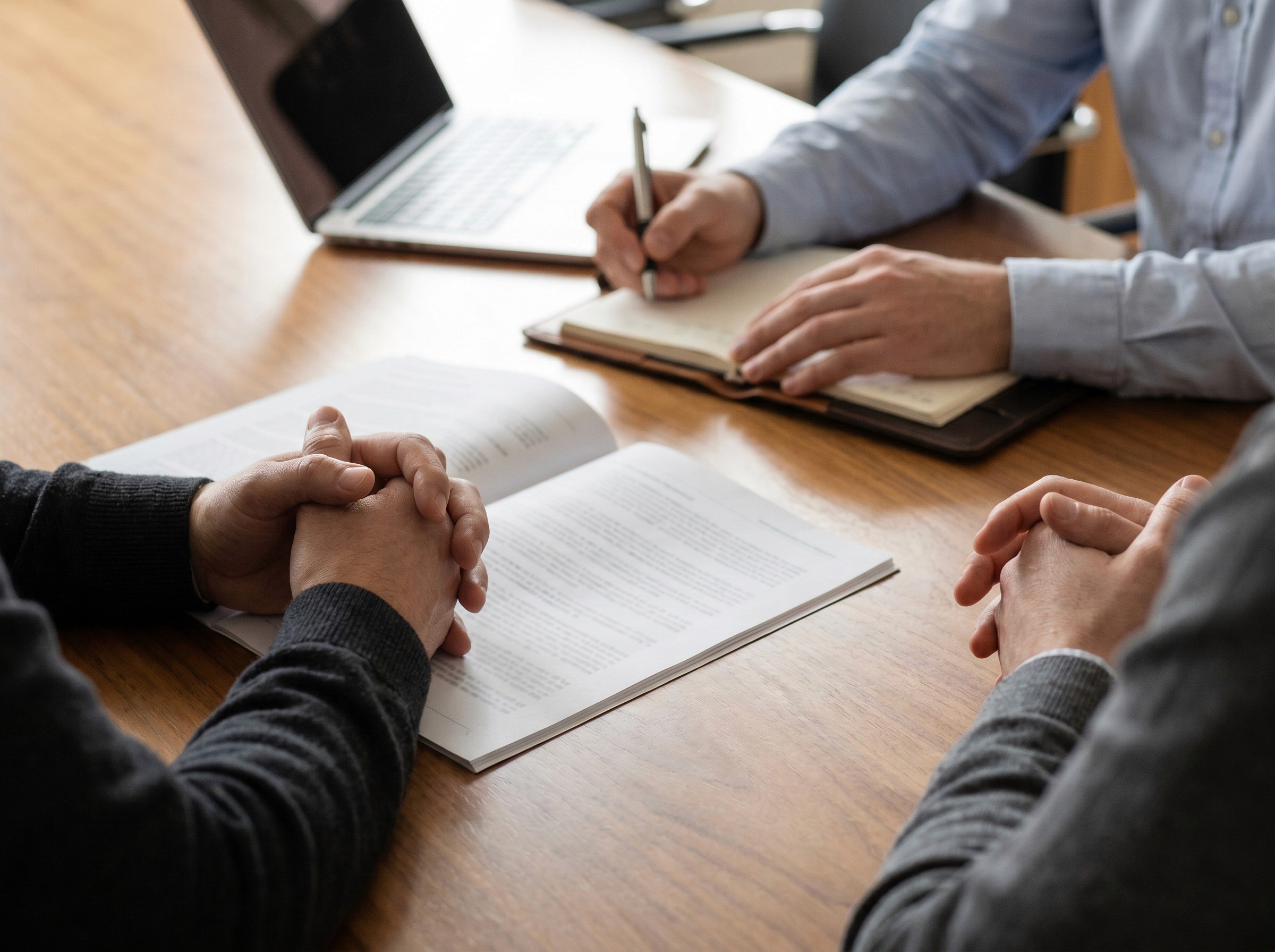 A close-up of hands at a boardroom table during a meeting. One person's hands are resting on a printed report, another person's hands are taking notes. A laptop screen is partially visible but blurred in the background. The focus is on the physical act of governance: reviewing, noting, deciding. Neutral colour palette, warm timber table surface. Shallow depth of field on the hands and document. No faces visible. No logos, no readable text on the report. Documentary style.