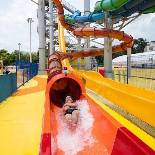 Man sliding down a bright red water slide at a water park, with colorful slides in the background and water splashing around him.