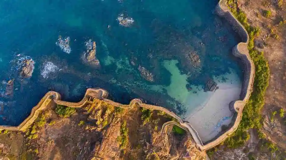 <img src="investment-in-sindhudurg-port.webp" alt="Aerial view of a historic coastal fort with stone walls curving along the rocky shoreline and enclosing a small sandy beach, with clear blue-green sea water and scattered rocks visible offshore.">