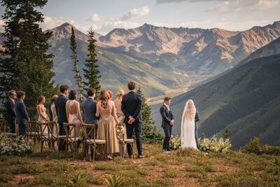 Guests stand beside wooden chairs as the couple face each other above the mountain valley.