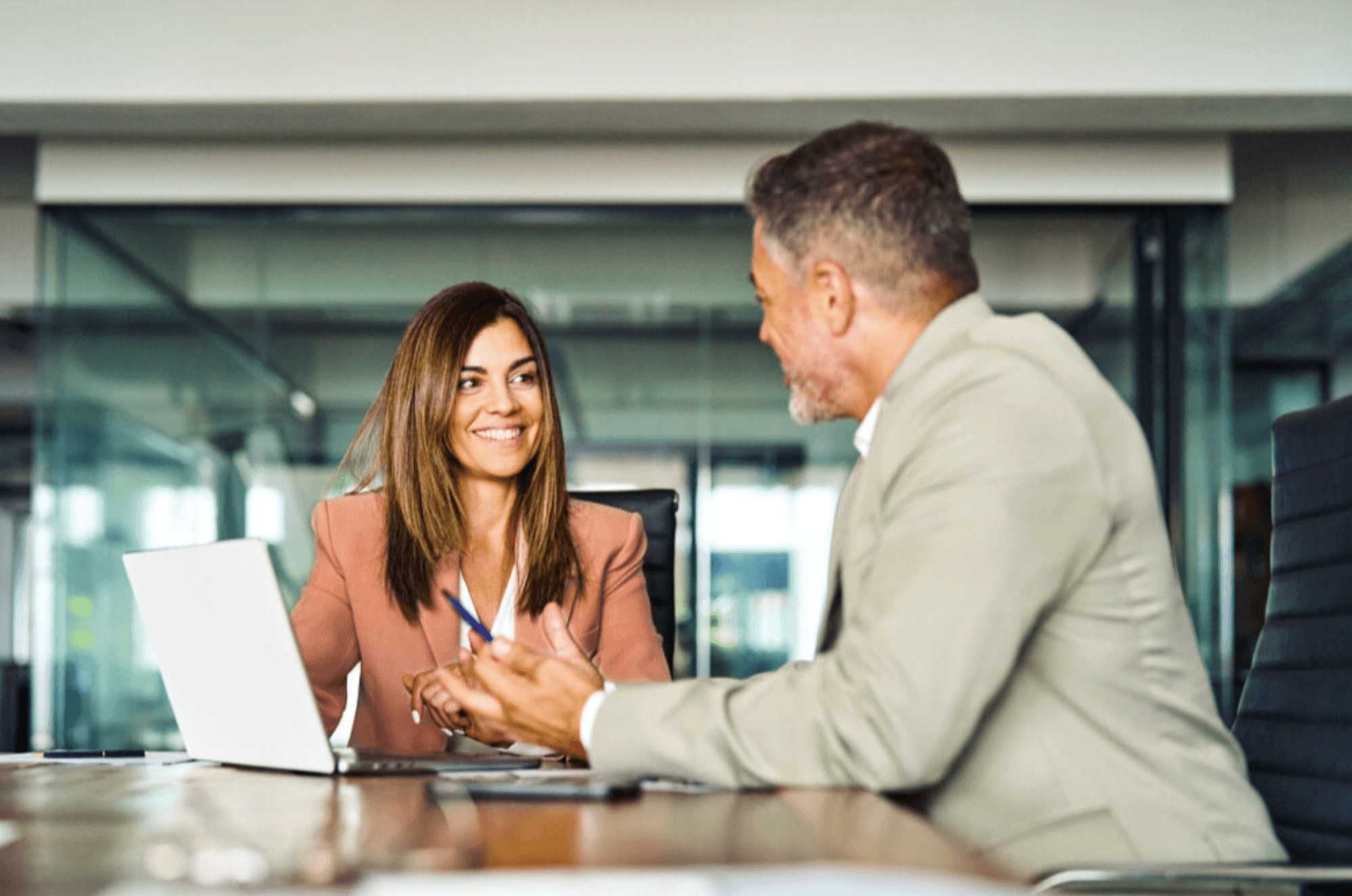 A woman in a pink blazer smiles while looking at a man in a light suit during a discussion at a conference table with a laptop. The professional setting features glass office walls in the background.