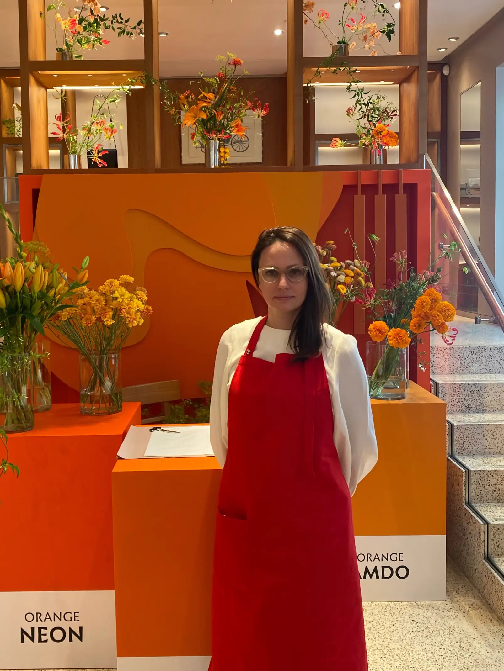 Irina Khatsernova con delantal rojo y blusa blanca posando frente a una instalación floral en tonos naranjas y amarillos. El fondo presenta estructuras geométricas color naranja con arreglos florales vibrantes en jarrones de vidrio. La escena transmite una estética cálida, moderna y artística inspirada en la energía del color naranja.