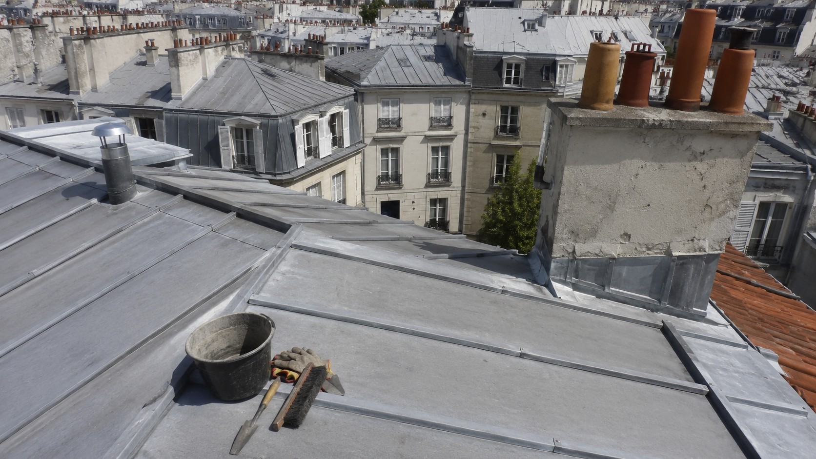 Toiture parisienne en zinc sous le soleil avec outils d’entretien, cheminées et immeubles haussmanniens, illustrant la préparation d’une toiture pour l’été à Paris.