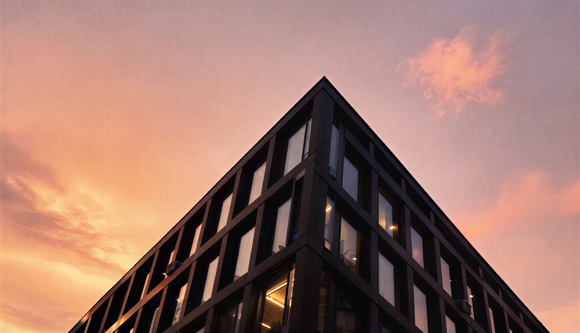 A low-angle view looking up at the sharp corner of a dark, modern gridded office building against a vibrant orange and pink sunset sky.