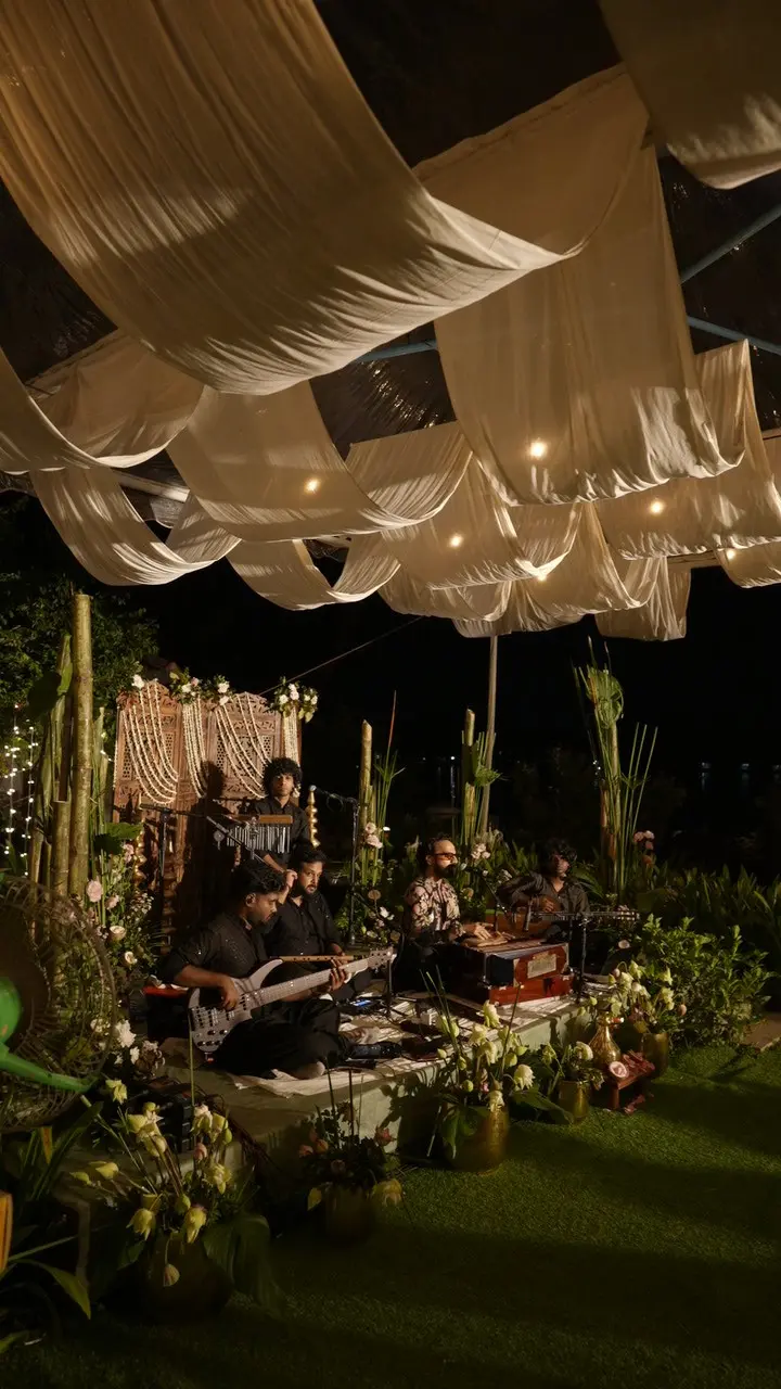 Live musicians performing under white fabric canopy at a Vedic Village Alleppey evening event, with fairy lights, floral arrangements and heritage carved backdrop