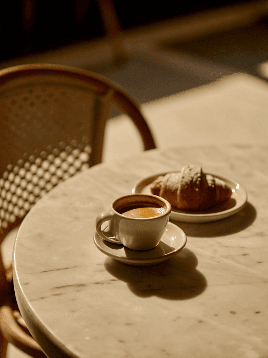A warm cappuccino in a white cup and a croissant on a plate rest on a sunlit marble table, exuding a cozy and inviting morning ambiance.