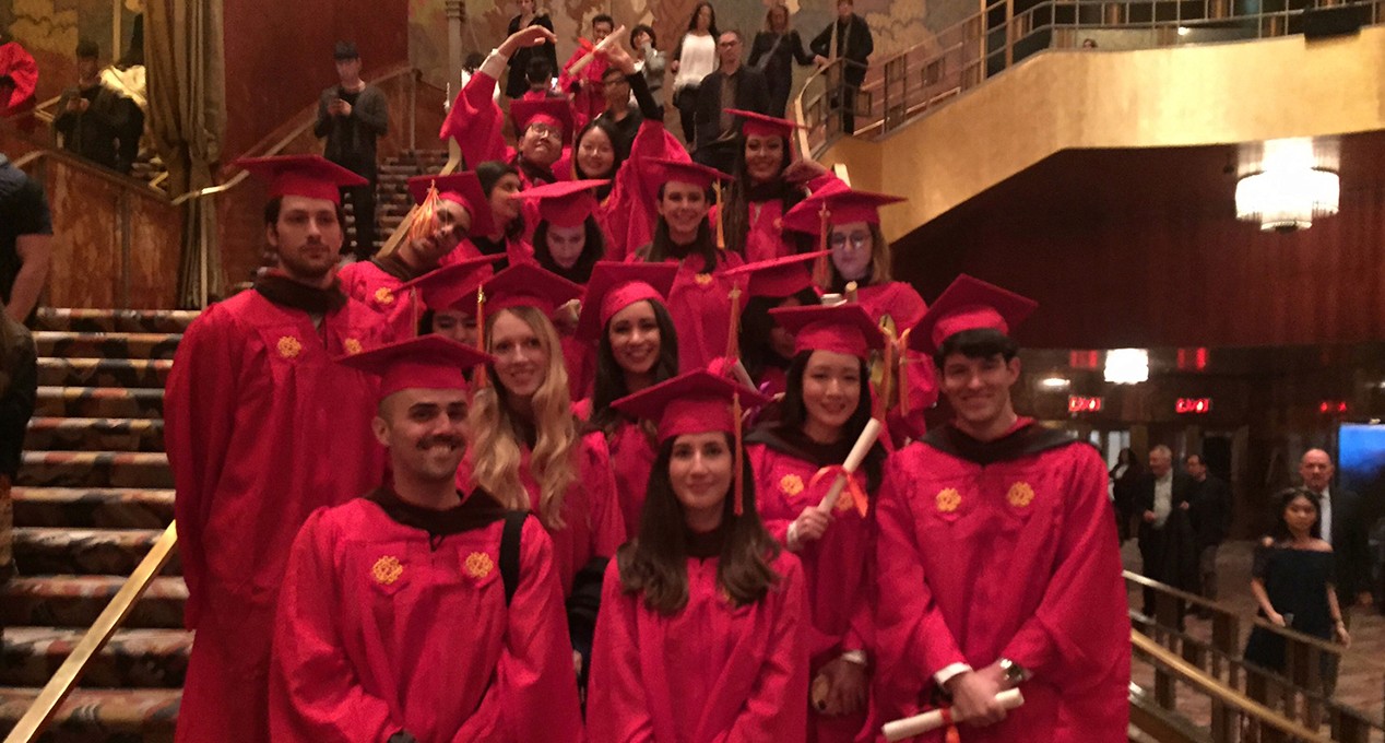 A photo of a couple of grad students dressed in SVA color robes, that are standing on the stairs of a festive hall.