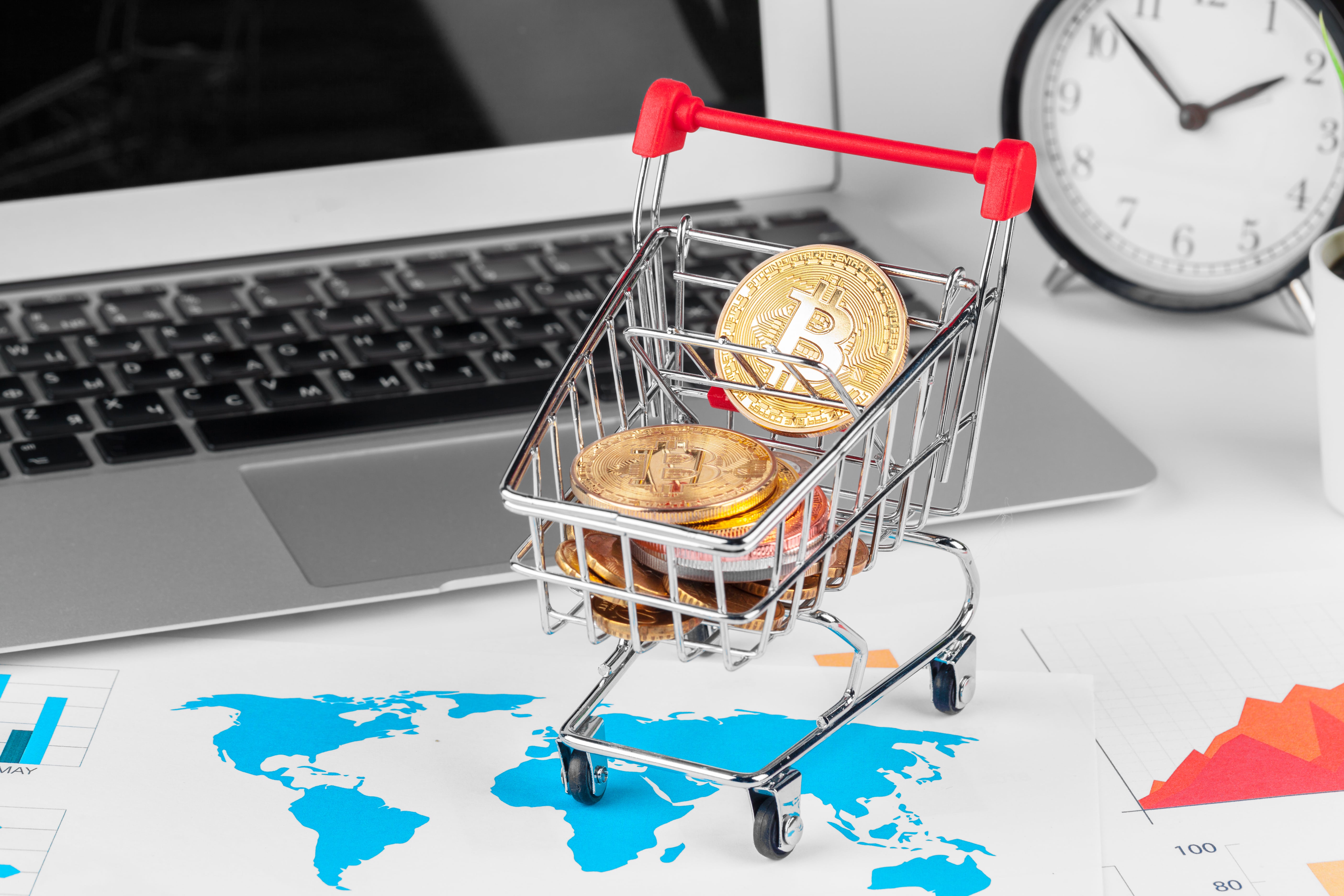 An image conceptualizing cryptocurrency investment and e-commerce, featuring a miniature shopping cart filled with golden Bitcoin coins. The cart is placed on an office desk on top of a world map and financial charts, with a laptop and an alarm clock in the background, symbolizing the fast-paced, global nature of online crypto trading.