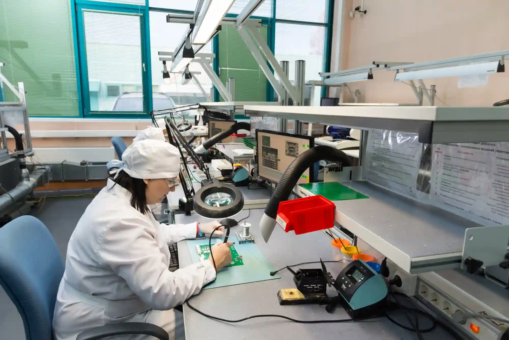 A technician in a sterile lab assembly environment carefully soldering components onto a green printed circuit board.