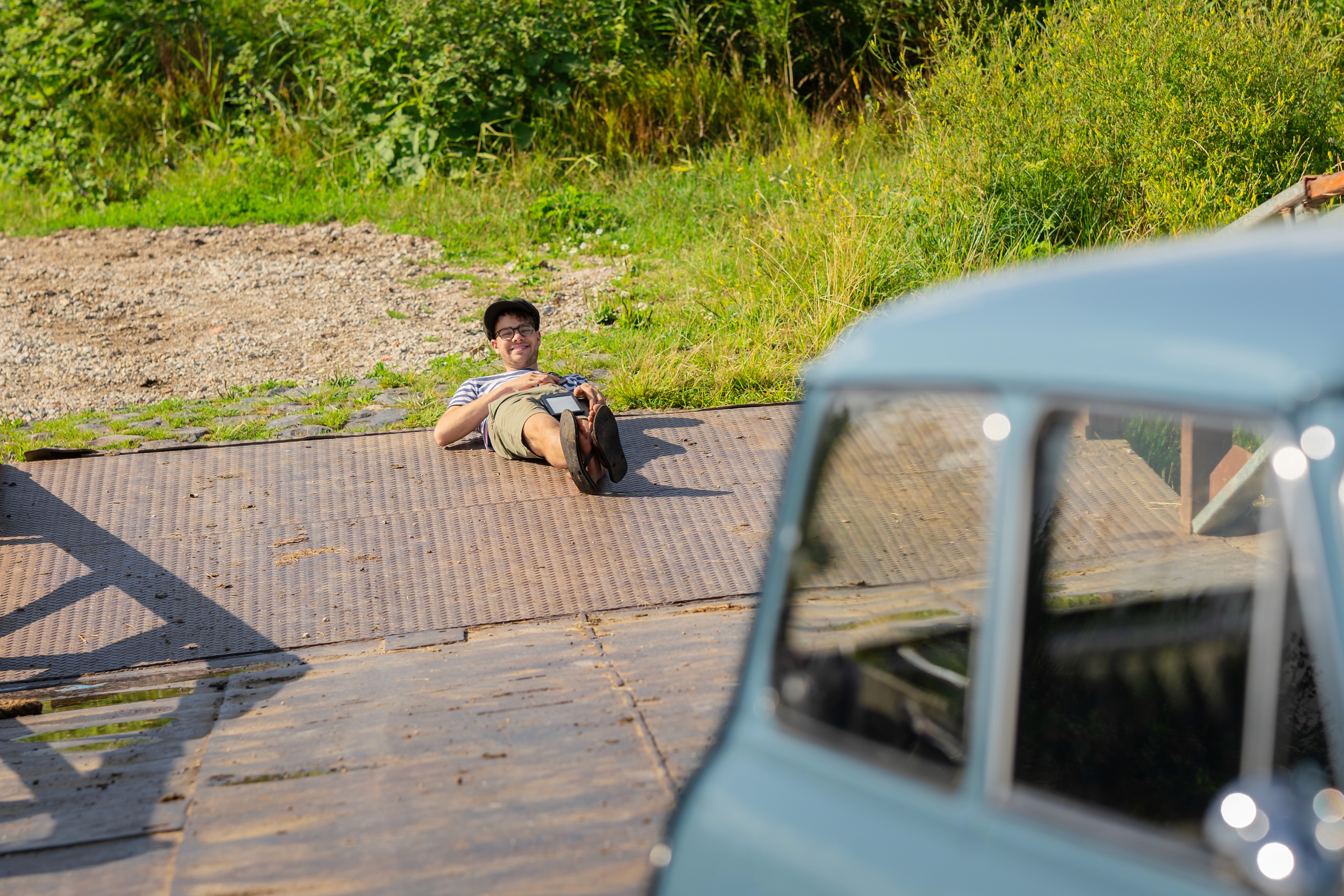 A person sits on the ground near a vintage car, surrounded by greenery and gravel. Sunlight illuminates the scene.