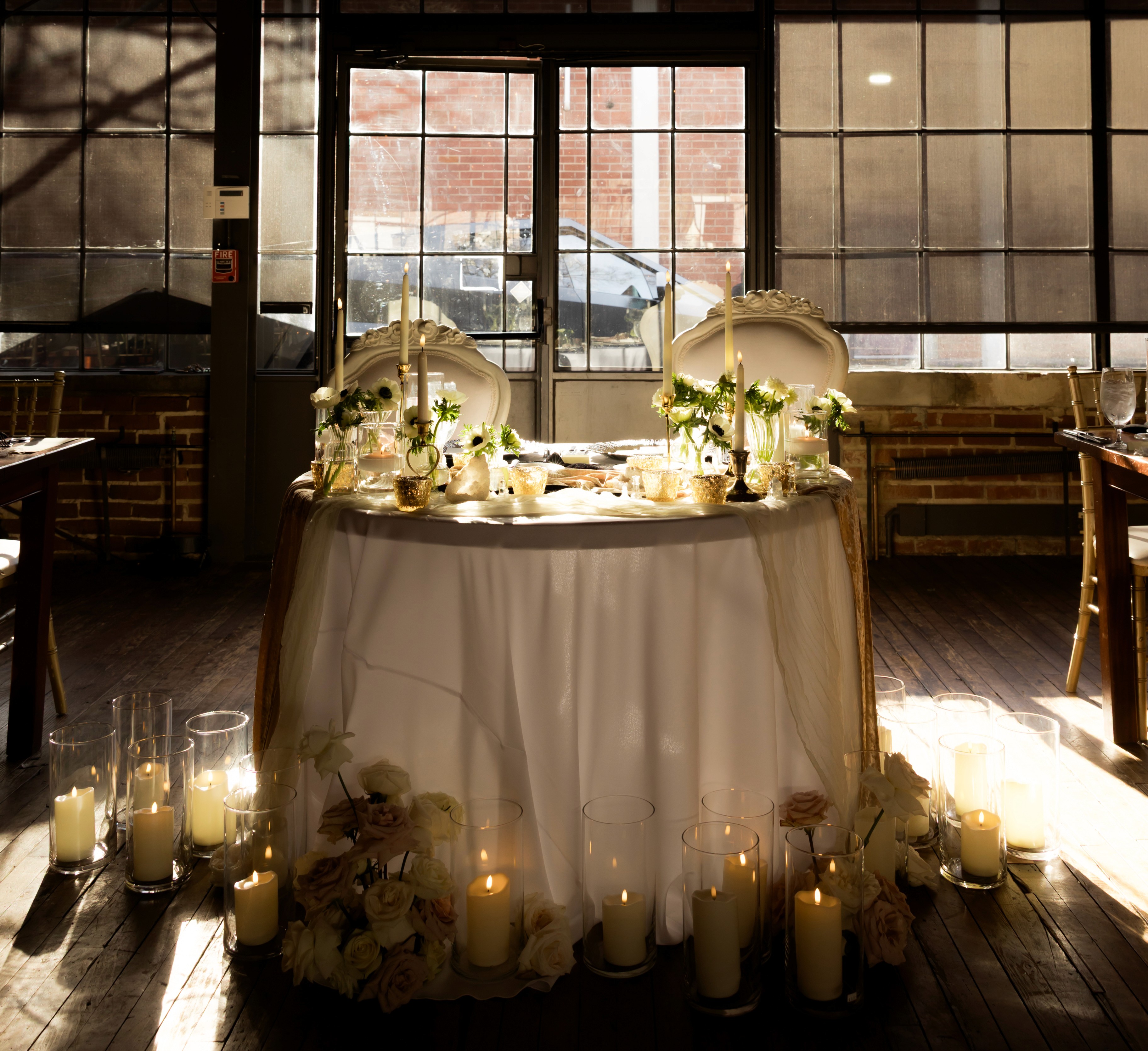 Candlelit reception table with white flowers, ornate chairs, and tall windows.