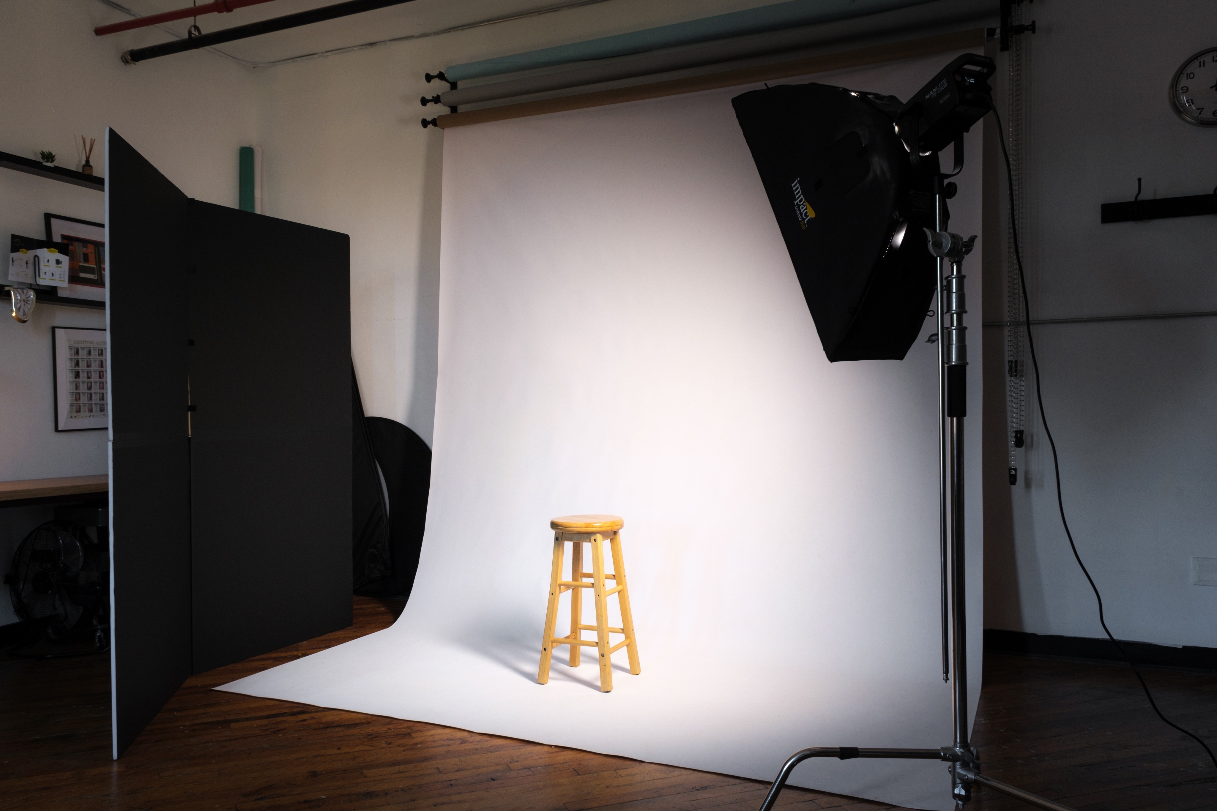 A daylight photography studio featuring a large wall of colorful backdrops, professional lighting equipment, and a workspace with shelves displaying decor and camera gear. A floral wall installation and a large mirror add artistic elements to the studio’s bright, versatile environment.