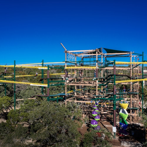 Large outdoor ropes course with multiple levels, obstacles, and climbers. Trees and a clear blue sky in the background.