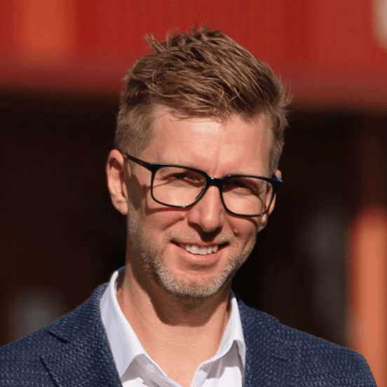 A person with glasses and a beard, wearing a navy polo shirt, sits in front of a modern red and black geometric building, smiling softly at the camera.